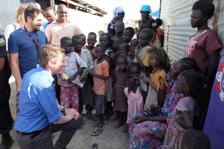 A group of adults and children gather outside a building in Sudan. One adult kneels, interacting with the children. Two uniformed UK peacekeepers, reminiscent of the efforts led by Williamson, stand nearby.
