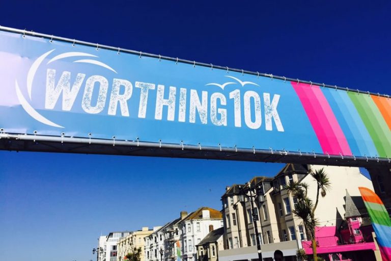 A banner reading "Worthing 10K" with colorful stripes flutters heroically against the clear blue sky. In the background, buildings and palm trees stand proudly, creating a vibrant backdrop that complements the spirited event.