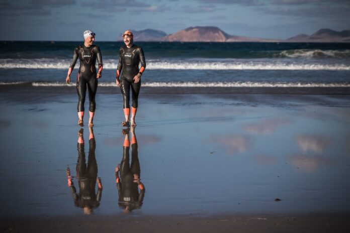 Two athletes in wetsuits and swim caps stroll along the beach, preparing for a Victory Swims triathlon. The ocean waves crash behind them, with distant mountains completing the breathtaking backdrop.