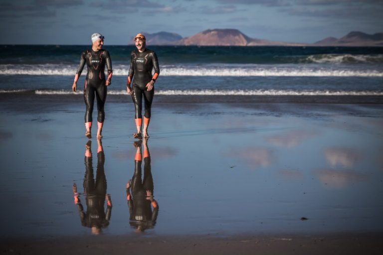 Two athletes in wetsuits and swim caps stroll along the beach, preparing for a Victory Swims triathlon. The ocean waves crash behind them, with distant mountains completing the breathtaking backdrop.
