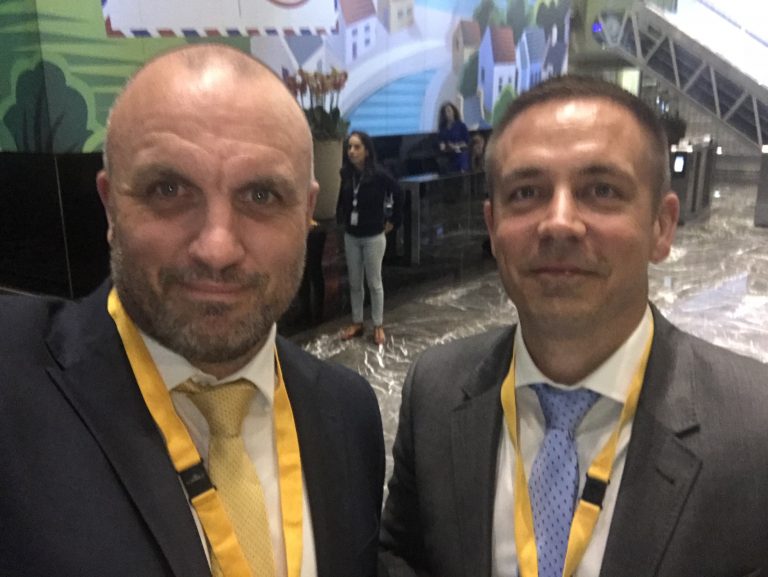 Two men in suits and yellow lanyards stand side by side in a large indoor space with a mural and person in the background, capturing the spirit of Cyber Pathways Insight Day.