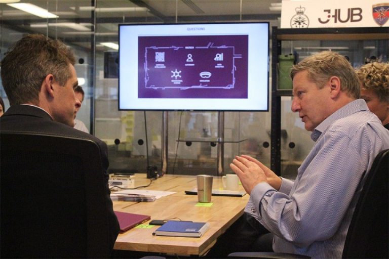 Two men are in a meeting room discussing, with a presentation on jHub displayed on a screen in the background.