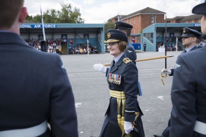 A three-star female commander walks in a parade ground, wearing a ceremonial sword and medals, while others in similar uniforms stand nearby. A crowd watches from stands labeled 