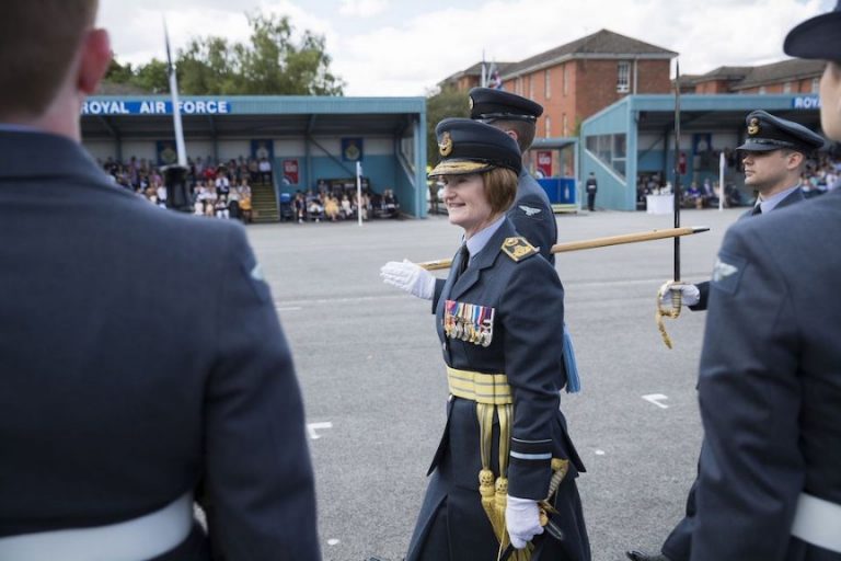 A three-star female commander walks in a parade ground, wearing a ceremonial sword and medals, while others in similar uniforms stand nearby. A crowd watches from stands labeled "Royal Air Force.