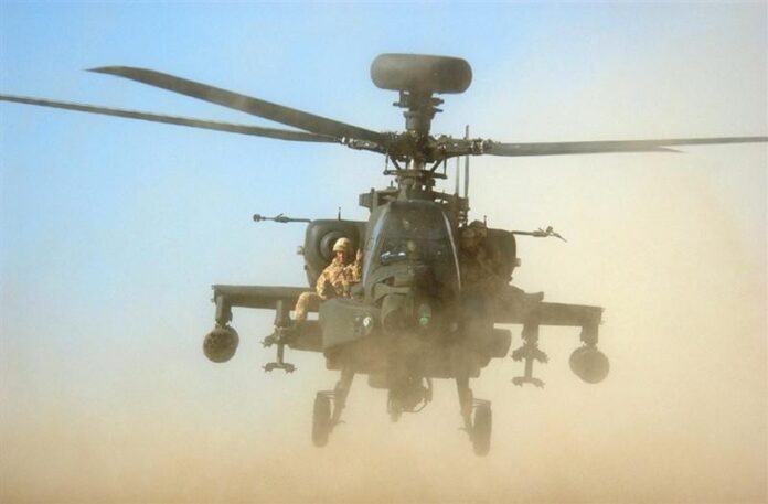 An army chopper flies low over a dusty terrain, preparing to touch down with armed personnel on board.
