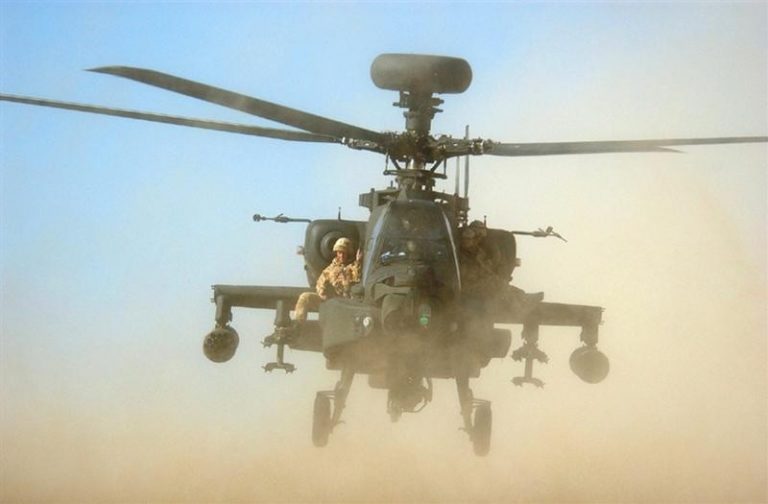 An army chopper flies low over a dusty terrain, preparing to touch down with armed personnel on board.