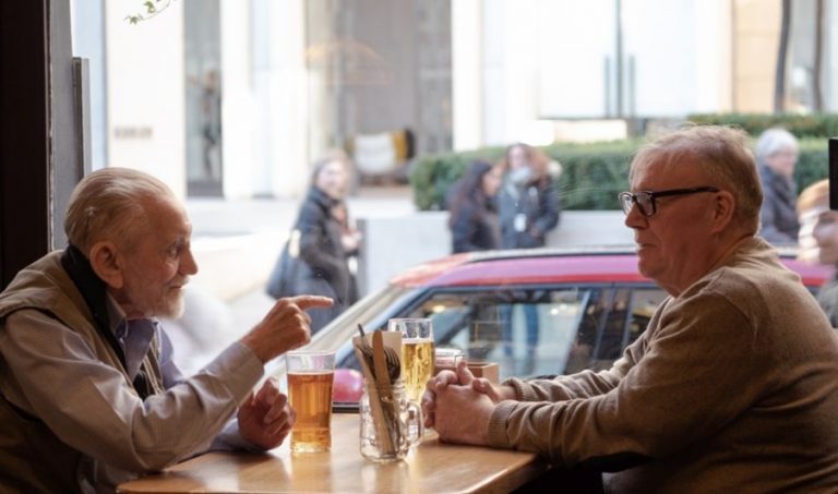 Two people sit at a table by a window, immersed in conversation. They each have a drink. Outside, the scene is like film highlights—people and cars blur past. Despite the busy street, there’s an aura of isolation around them as they share this moment away from the world’s noise.