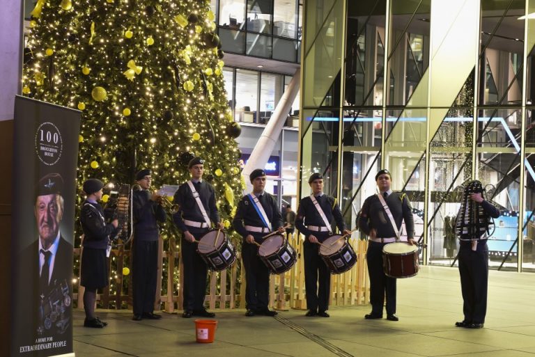 A group of uniformed individuals performs in front of a large, decorated Christmas tree near a modern glass building, symbolizing the expand forces initiative. A sign with a portrait is proudly displayed beside them.