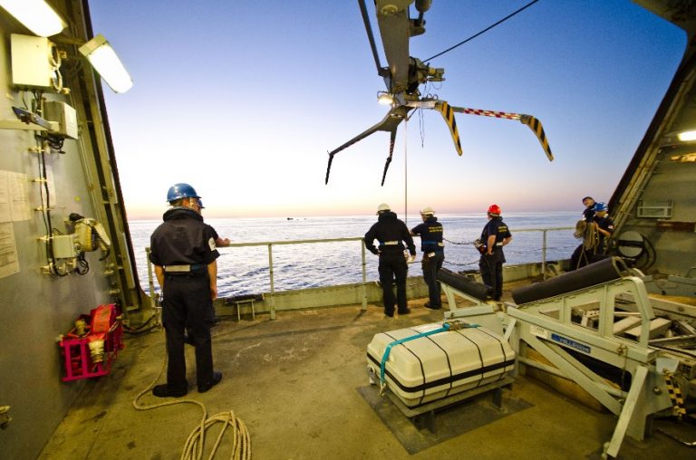 Crew members in helmets stand on the deck of HMS Dragon, glancing at the sea during a stunning sunset, their minds perhaps reflecting on their recent successful drugs haul in the Middle East.