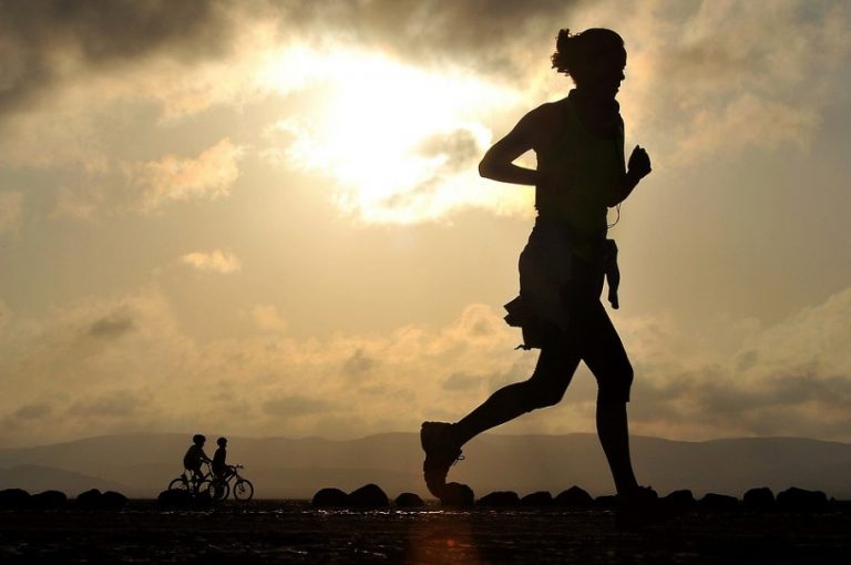 Silhouette of a person running in the foreground, perhaps training for a marathon challenge, with the sun shining behind and two cyclists in the background.