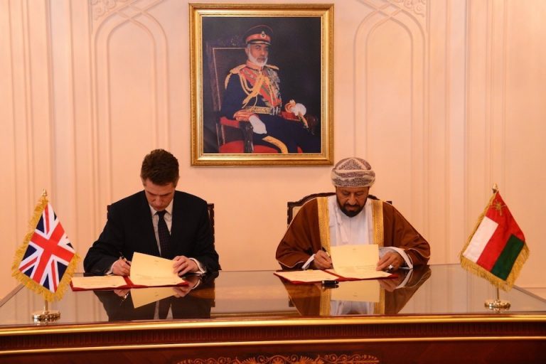 Two men seated at a table sign documents related to a Defence Agreement, with the UK and Oman flags displayed prominently. A portrait hangs on the wall behind them.