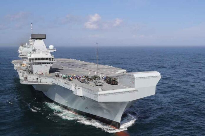 An aircraft carrier, a testament to Global Britain's strength, sails through the ocean under a clear sky.