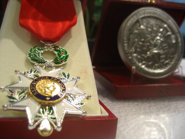 Close-up of a Légion d’Honneur medal in a box, accompanied by a round silver medal in the background. These prestigious honors, often awarded to veterans of D-Day, represent bravery and heroism.