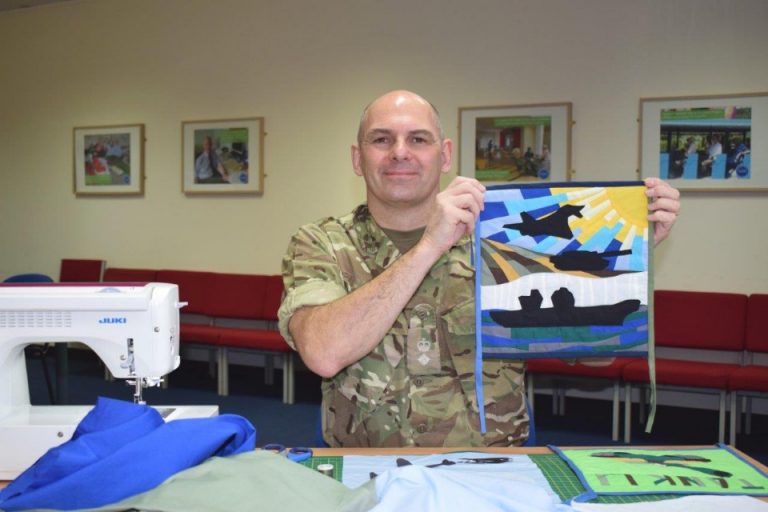 A sewing soldier in military uniform proudly holds up a colorful quilt piece with a silhouette design. With gratitude for his craft, flags inspire the vibrant patterns. A sewing machine and fabric are spread across the table in front of him, showcasing his dedication to art and service.