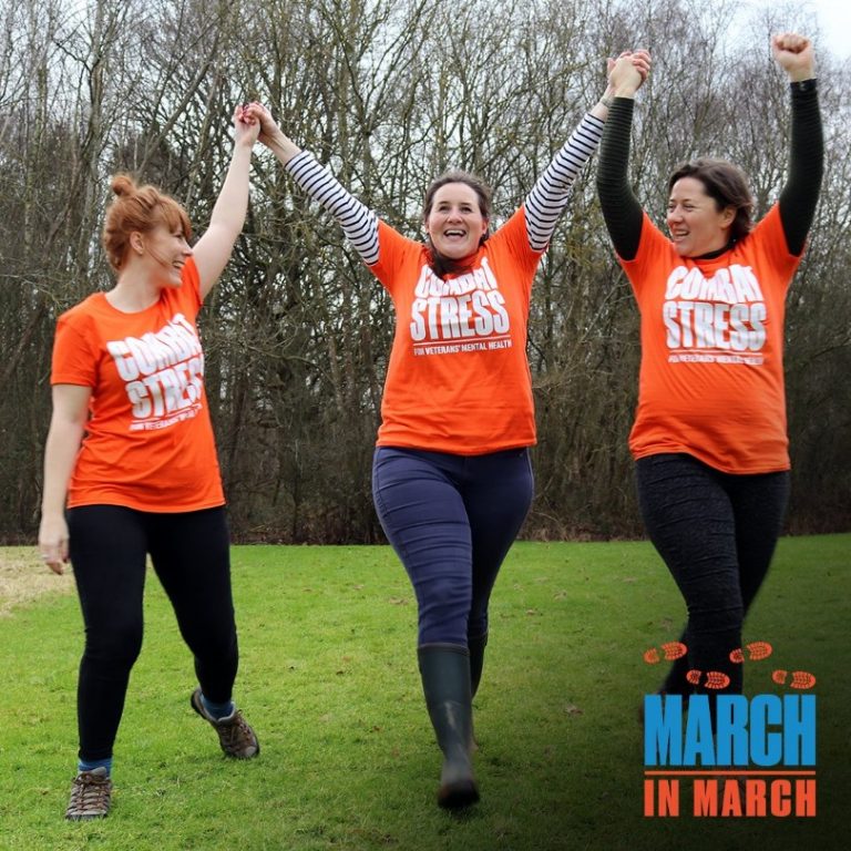 Three women in orange "Combat Stress" shirts hold hands and smile as they walk outdoors on grass, embodying the spirit of "Boot Forward." Trees provide a lush backdrop, while the "March in March" logo sits proudly in the bottom right corner.