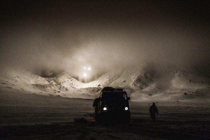 A truck and a Marine navigate the snowy, mountainous landscape at night, under the ethereal glow of the Arctic night sky, with bright lights cutting through the fog.