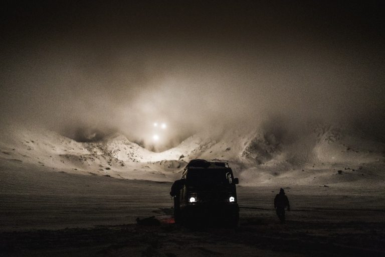A truck and a Marine navigate the snowy, mountainous landscape at night, under the ethereal glow of the Arctic night sky, with bright lights cutting through the fog.
