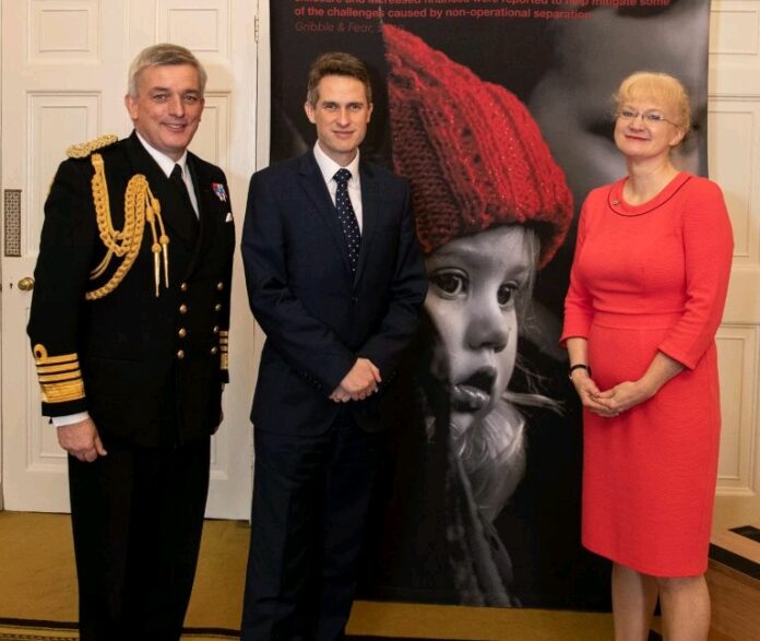 Three people stand in front of a poster featuring a child. The person on the left is in a military uniform, suggesting the complexities of family separation, the person in the center is in a suit, and the person on the right radiates confidence in a red dress.