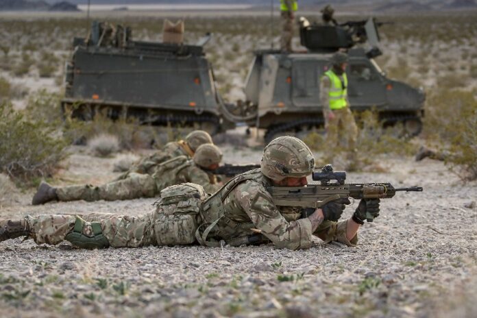 Royal Marines in camouflage uniforms lie on the ground, aiming rifles as armored vehicles loom in the desert landscape, honing their skills for Arctic Defence scenarios.