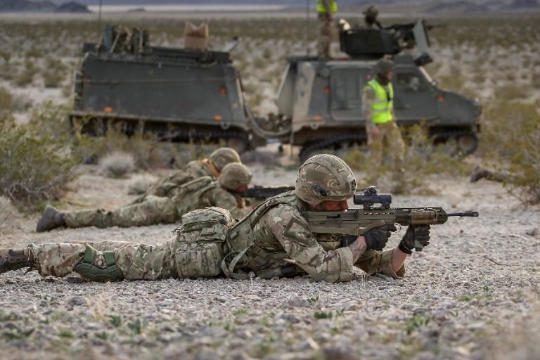 Royal Marines in camouflage uniforms lie on the ground, aiming rifles as armored vehicles loom in the desert landscape, honing their skills for Arctic Defence scenarios.