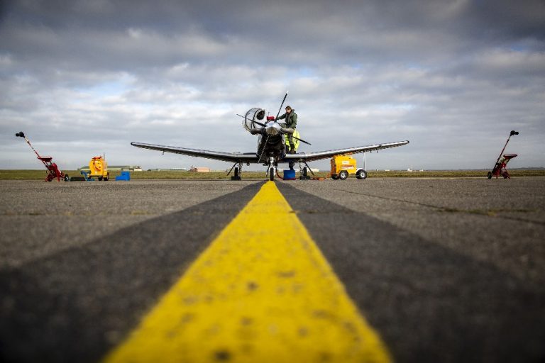 A small aircraft on a runway is undergoing maintenance, with a person diligently working to ensure everything is in top shape for aviation purposes. Support vehicles and equipment surround the scene under a cloudy sky, setting the stage for flight training that will take pilots to new heights.