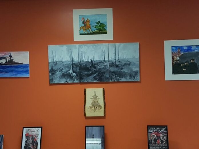 A variety of paintings and framed pictures, supported by Council Funding, are displayed on an orange wall.