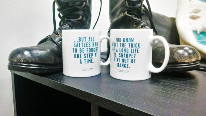 Two ceramic mugs with text sit on a black table, symbolizing camaraderie and teamwork, in front of a pair of worn black combat boots.