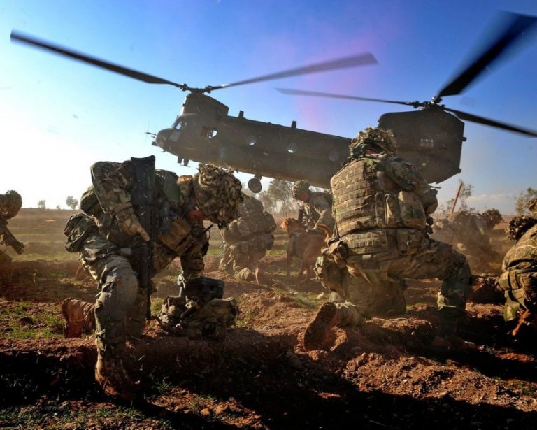 Soldiers in camouflage gear, part of the UK troops, crouch as a military helicopter lands in a dusty field.
