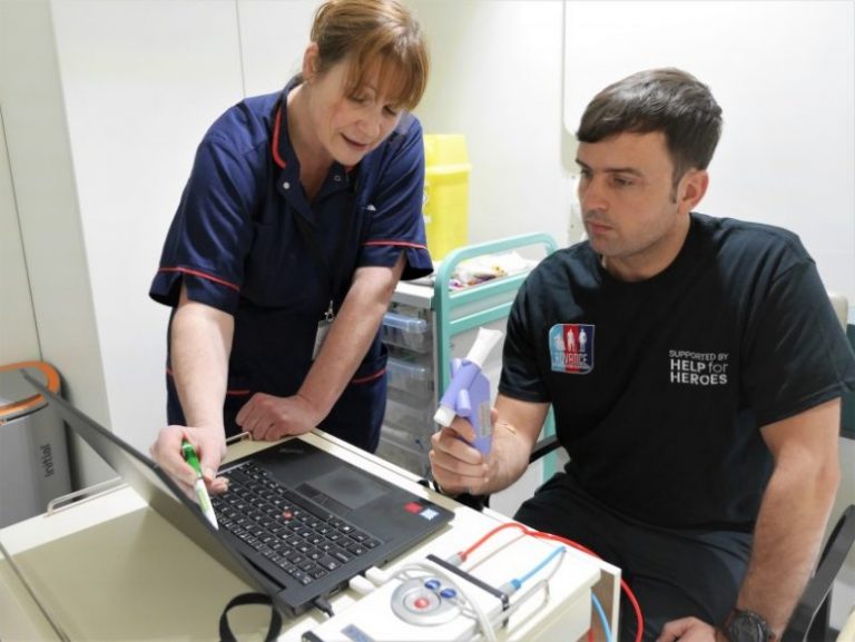 A nurse and a patient are intently uncovering information on a laptop screen. The patient, holding a medical device, seems engaged, while various medical equipment is neatly arranged on a cart in the background.