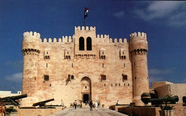 This historic stone fortress, featuring cylindrical towers and arches, stands as a testament to UK-Egypt cooperation in fortified architecture. A flag waves proudly above the central entrance, where cannons are strategically placed under a partly cloudy sky.