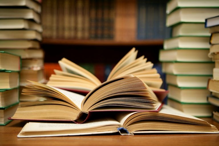 Open books stacked on a wooden table, surrounded by towering piles of closed books in a library setting—a perfect scene to celebrate World Book Day.