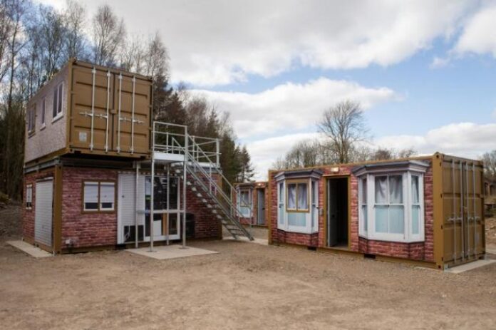 Container homes in a rural setting near Catterick, with stairs leading to a second level, surrounded by trees under a cloudy sky.