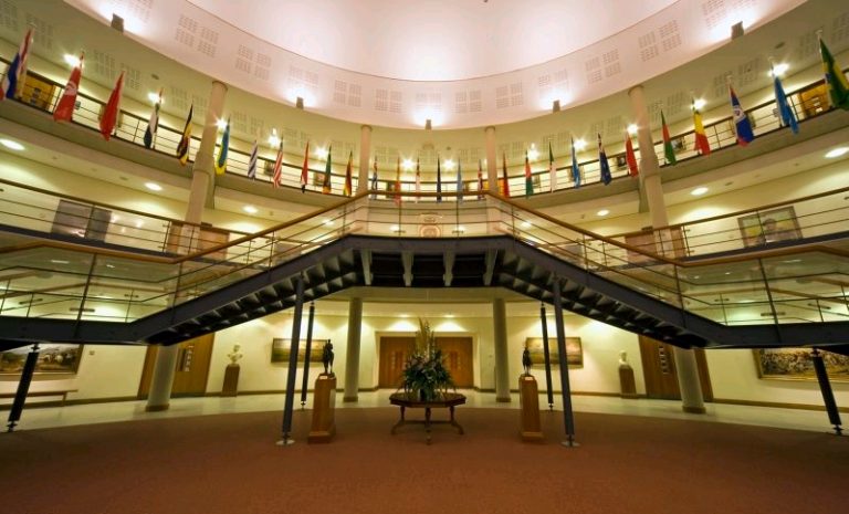 A grand, circular lobby with a dual staircase, flags adorning the upper level, and a central plant arrangement showcases modern security.