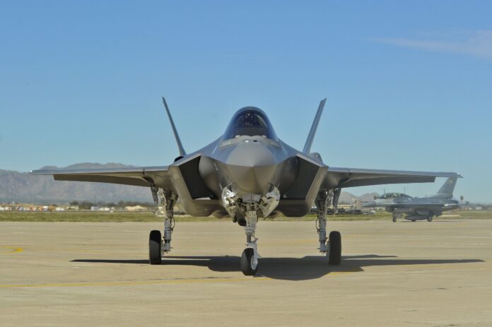 An F35 jet is parked on a runway at Lakenheath, with towering mountains in the background. Another aircraft is visible in the distance, marking its arrival.