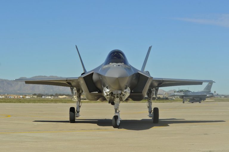 An F35 jet is parked on a runway at Lakenheath, with towering mountains in the background. Another aircraft is visible in the distance, marking its arrival.