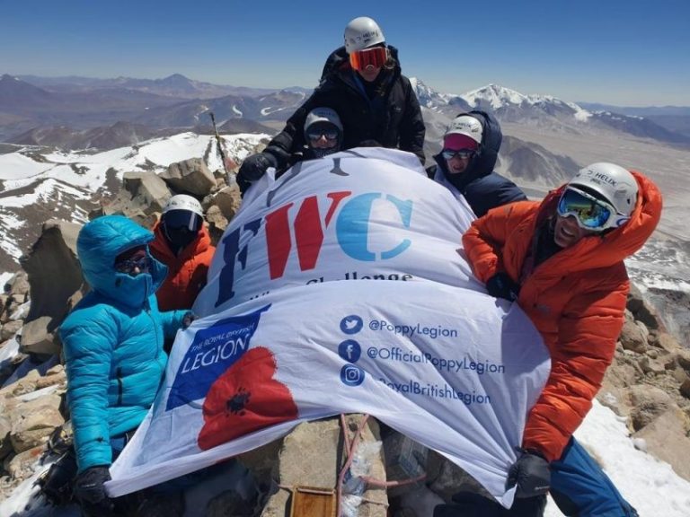 A group of climbers in winter gear proudly holds an "FWC" banner amidst the majestic peaks, having just conquered the summit of the world's highest volcano.