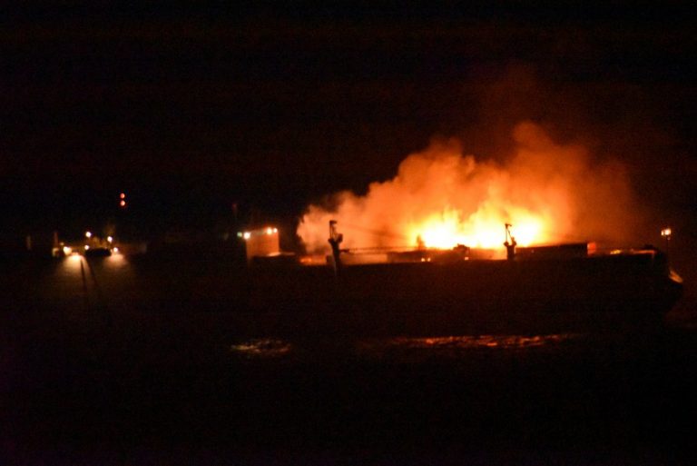 A blazing ship illuminates the night as bright flames and smoke rise into the dark sky. Nearby lights spotlight parts of the vessel, while HMS Argyll swiftly approaches to rescue the crew from the inferno.