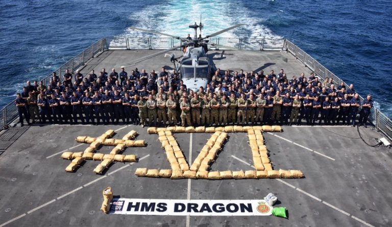A large group of military personnel stands in formation on the deck of a destroyer ship, surrounding a helicopter and arranged next to "#VI" and "HMS DRAGON," ready to deal a blow to terrorists threatening peace.