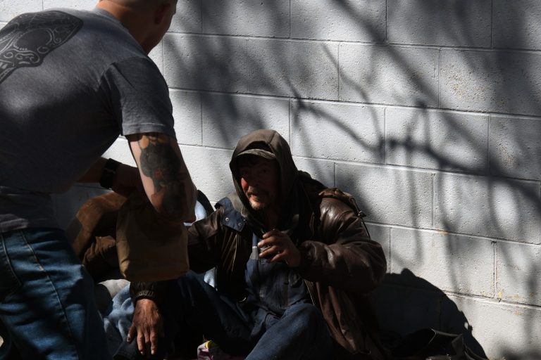 A person hands a paper bag to a homeless veteran sitting on the ground against a wall, partially in shadow.