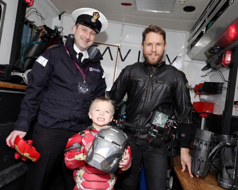 A man in a navy uniform, alongside his son—a smiling child dressed as Iron Man—pose with a man in a leather jacket. The child proudly holds a silver mask inside the truck's interior.