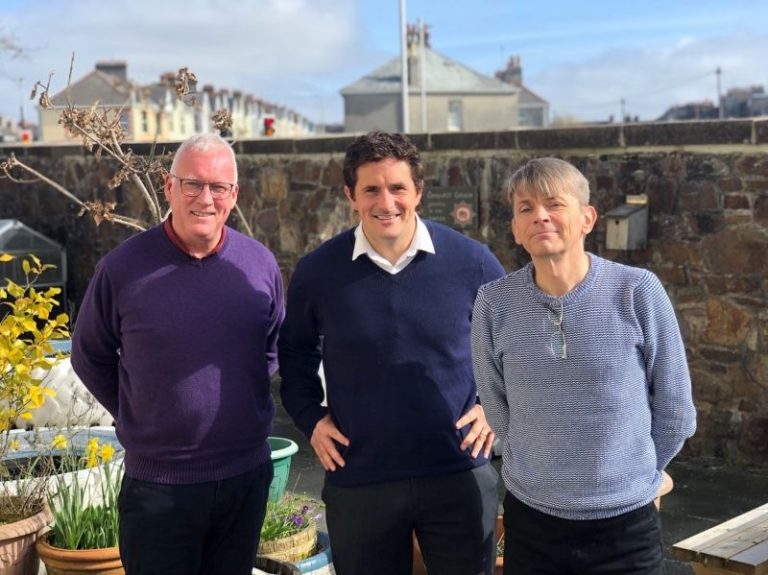 Three men, all veterans, stand outdoors with smiles on their faces, a row of houses behind them. The sky is partly cloudy.