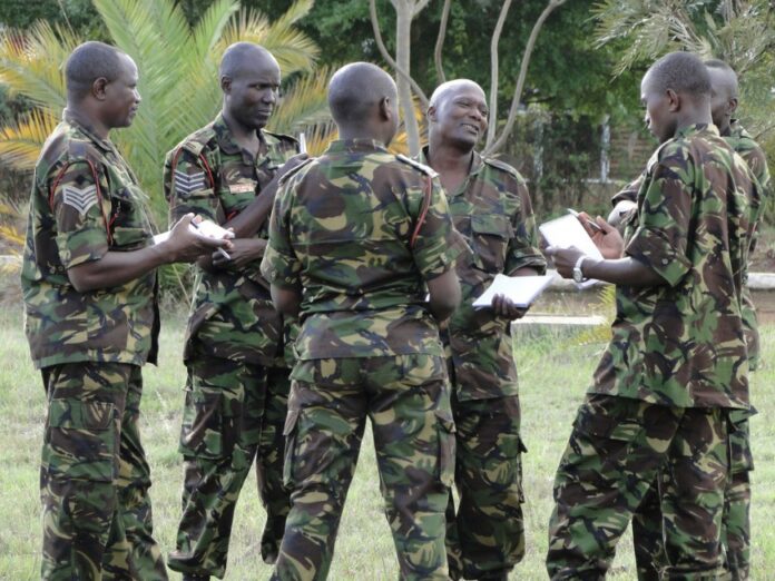 A group of soldiers in camouflage uniforms stands in a circle outdoors, engaged in discussion about the UK-Kenya Security Dialogue while holding notebooks.