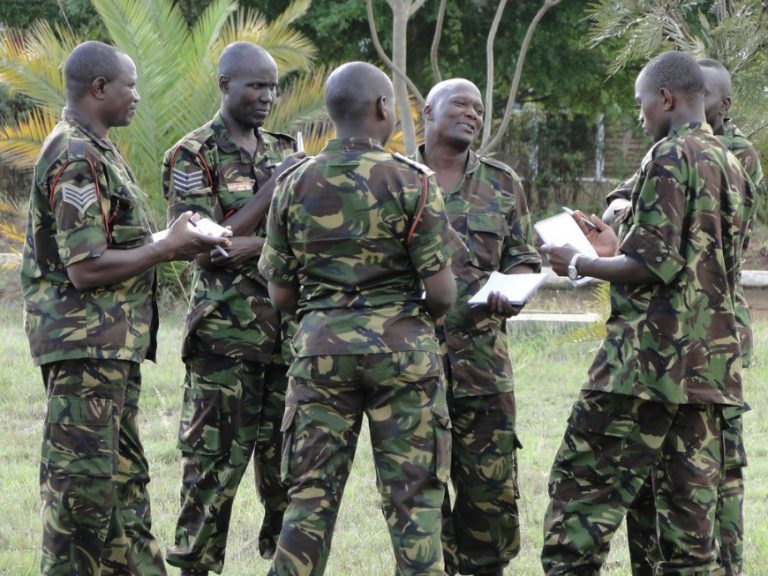 A group of soldiers in camouflage uniforms stands in a circle outdoors, engaged in discussion about the UK-Kenya Security Dialogue while holding notebooks.