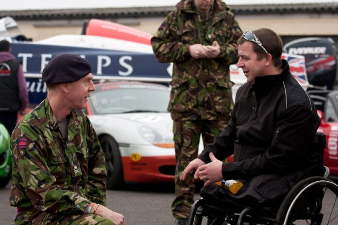 A man in a wheelchair talks to a man in a military uniform at an event with race cars in the background, discussing a newly launched initiative aimed at boosting veterans' employment.