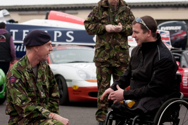 A man in a wheelchair talks to a man in a military uniform at an event with race cars in the background, discussing a newly launched initiative aimed at boosting veterans' employment.