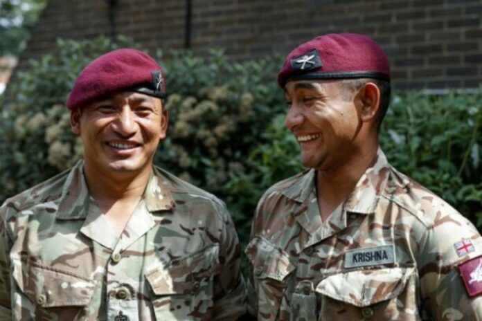 Two soldiers in camouflage uniforms and maroon berets smile while standing outside, representing the New Gurkha Battalion against a backdrop of greenery and a brick wall.