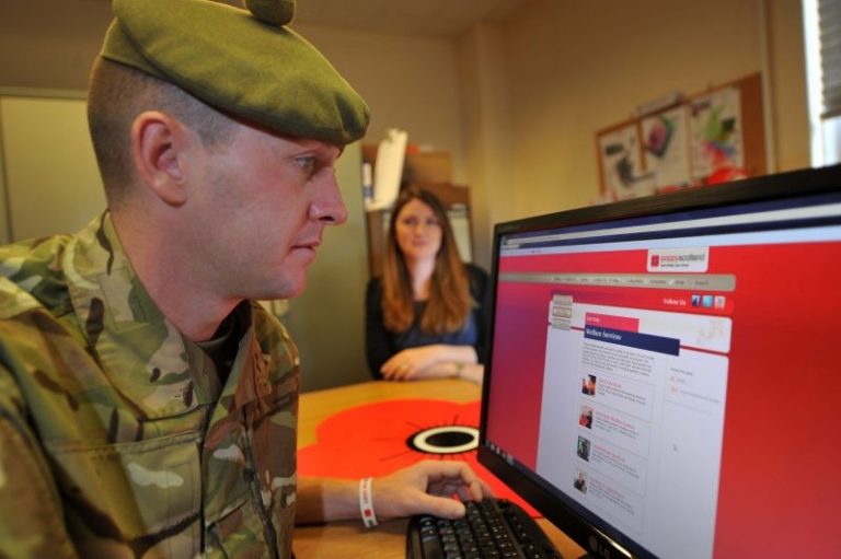 A person in military uniform uses a computer with a woman in the background, facing a screen displaying Poppyscotland's website with red and white colors, preparing for an upcoming Employment Open Day.