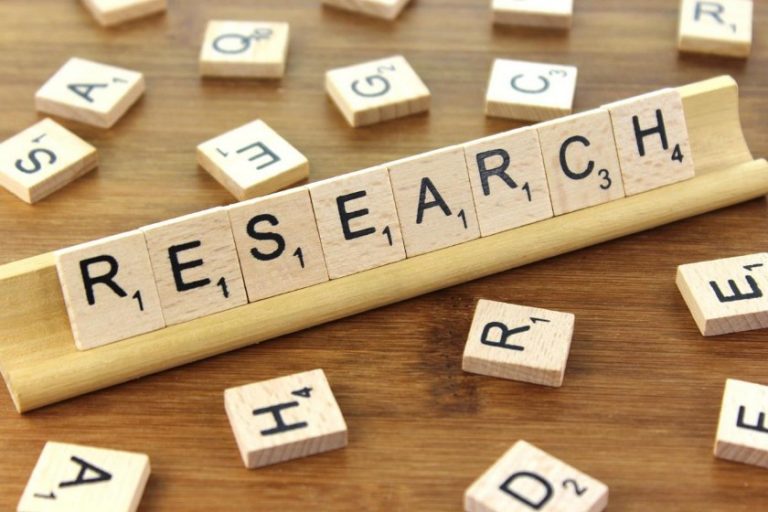Scrabble tiles spelling "RESEARCH" are artistically arranged on a wooden rack, with scattered tiles on the surface hinting at an intense study in progress.