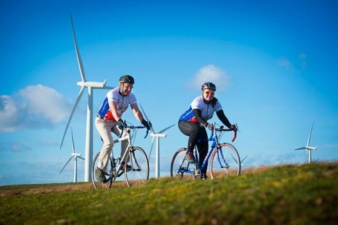Two cyclists ride like the wind on a grassy path that weaves through a serene wind farm, with several towering turbines spinning against the backdrop of a clear blue sky.