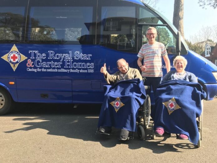 Two people in wheelchairs with blue blankets are accompanied by a man standing behind them, posing in front of a blue bus labeled 